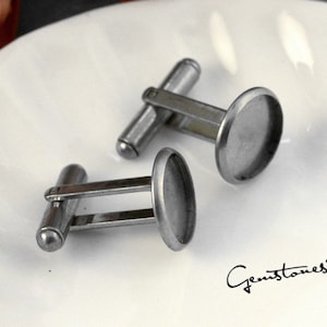 Puede incluir: Un par de gemelos plateados con un diseño clásico. Cada gemelo presenta una barra redondeada y una cara plana de forma ovalada. Los gemelos se muestran sobre una superficie blanca. La imagen incluye el texto "Gemstones2u" en cursiva.