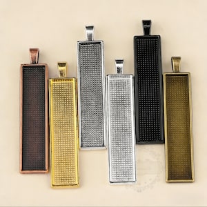 Puede incluir: Una colección de bandejas rectangulares para colgantes en varios acabados metálicos. Las bandejas son de bronce, oro, plata y negro. Cada bandeja tiene un interior texturizado y un pequeño lazo en la parte superior para sujetar una cadena o cordón.