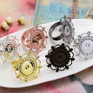 May include: Six ornate metal ring blanks with openwork designs and round bezels. The bezels are different colors: gold, silver, bronze, and black.