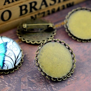 May include: Two antique bronze-coloured metal settings for glass cabochons. One setting has a white background with a blue butterfly design. The other setting is empty.