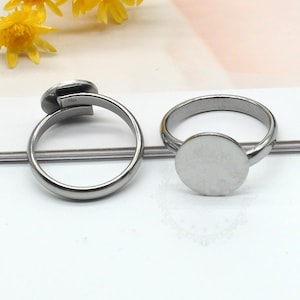 May include: Two silver rings. One ring has a small, oval setting, the other a circular setting. The rings are on a white surface, with yellow flowers in the background. The rings are made of a silver-toned metal.