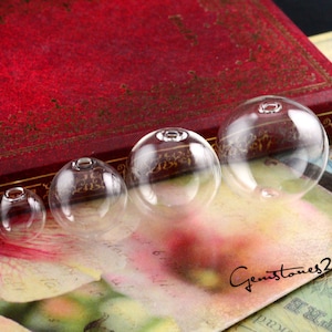 May include: Four clear glass beads of varying sizes, arranged from largest to smallest, on a vintage book and a patterned surface. The beads are smooth and round, with a small hole at the top.