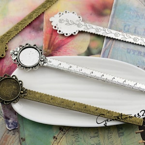 Puede incluir: Primer plano de varios colgantes metálicos en forma de regla con engastes florales y redondos. Las reglas son de tonos bronce antiguo y plata, con medidas grabadas y diseños florales. Ideal para la creación de joyas.