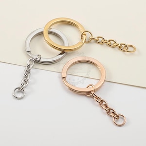 Op de afbeelding: Drie metalen sleutelhangers in goud, zilver en roségoud. Elke sleutelhanger heeft een grote cirkelvormige ring met een kleine ketting en een kleinere ring. De sleutelhangers worden gepresenteerd op een lichtgekleurd oppervlak.