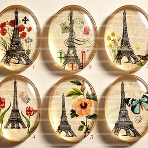 Op de afbeelding: Zes ovale glazen cabochons met zwarte illustraties van de Eiffeltoren op een beige textuurachtergrond. Elk stuk toont unieke accenten: rode bloemen, postzegels, gele bloemen met een vogel, rode tulpen, een oranje hibiscus en een blauwe vlinder.