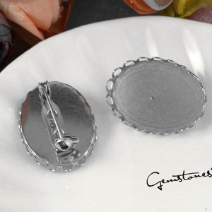 Op de afbeelding: Twee zilverkleurige ovale brochebases met decoratieve geschulpte randen. Eén basis heeft een speldrug bevestigd. De andere basis is leeg. De bases liggen op een wit oppervlak. De tekst "Gemstones2u" staat in de rechter benedenhoek.