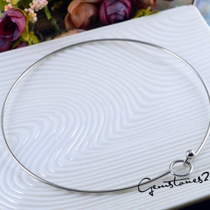 Pode incluir: Um colar choker prateado com um pequeno charme circular. O colar está sobre uma superfície branca com um padrão ondulado.