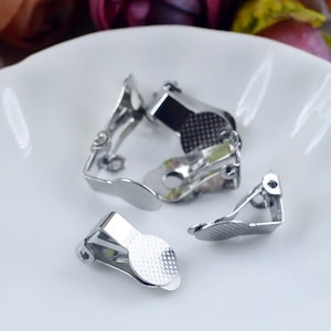 May include: Several silver-colored clip-on earring findings are arranged on a white, slightly scalloped plate. The earring findings have a simple, functional design with a textured, circular pad.