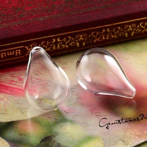 May include: Two clear, teardrop-shaped glass pendants. Each pendant has a small opening at the top and a rounded bottom. The pendants are transparent, allowing light to pass through. The background is a mix of colors, including red, pink, and green.