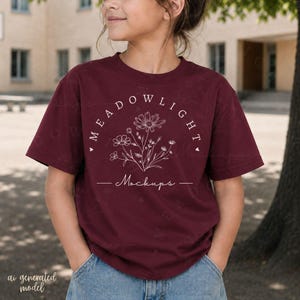 Può includere: Una t-shirt a maniche corte color bordeaux con una grafica a linee bianche raffigurante un bouquet di fiori di campo. Il testo MEADOWLIGHT è arcuato sopra i fiori, con la parola Mockups scritta in corsivo sotto il disegno floreale e cuori.
