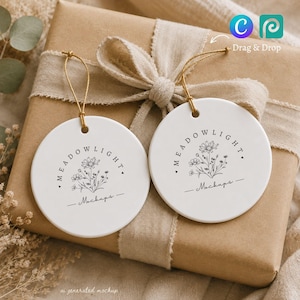 Può includere: Due ornamenti in ceramica bianchi di forma circolare con cordini dorati. Ogni ornamento presenta un'illustrazione a tratto nero di un bouquet di fiori di campo, il testo MEADOLIGHT e la parola Mockups in corsivo sotto i fiori.