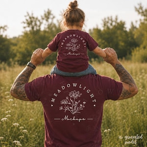 Può includere: Una t-shirt bordeaux indossata da un adulto e una t-shirt bordeaux più piccola abbinata indossata da un bambino. Entrambe le maglie presentano una grafica a linee bianche di un bouquet di fiori con il testo MEADOWLIGHT Mockups in cerchio.