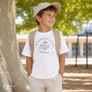 May include: A white short-sleeved t-shirt featuring a black line art illustration of a wildflower bouquet with the text "MEADOWLIGHT Mockups" printed on the front. The shirt is paired with a beige baseball cap and a beige backpack.
