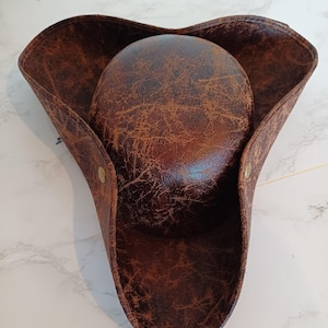 Peut inclure: Un chapeau tricorne avec une finition texturée marron foncé et vieillie. Le chapeau présente une calotte arrondie et un large bord relevé, fixé sur les côtés par de petites attaches métalliques circulaires, formant une silhouette triangulaire.