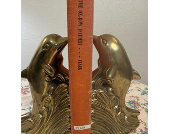 May include: A pair of polished brass dolphin bookends featuring a wave design at the base. Between the two dolphins stands a single orange hardcover book with the title The Ox-Bow Incident by Clark and the publisher Peter Smith on the spine.