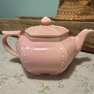 以下が含まれることがあります： 八角形の本体と蓋が付いた淡いピンク色の陶磁器製ティーポット。ティーポットには、湾曲したハンドル、先細りの注ぎ口、縁と蓋に沿ったロープのような装飾的なエンボス加工が施されています。光沢のある仕上げです。