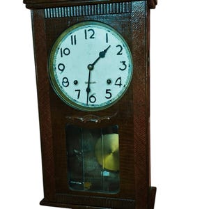 May include: A dark brown wooden wall clock with a rectangular frame and a decorative top crest. The white circular clock face features black Arabic numerals and two black hands. A glass panel below reveals a circular brass pendulum inside.
