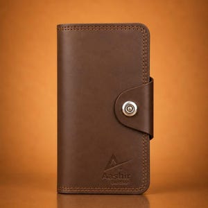 May include: A vertical brown leather wallet featuring a snap button closure on the side. The wallet has visible stitching along the edges and the embossed text Aashir Leather with a triangular logo symbol located at the bottom front center.