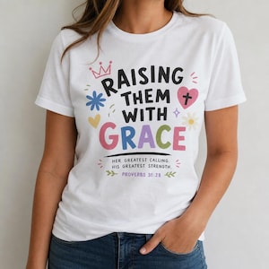 Puede incluir: Una camiseta blanca de manga corta con el texto negro RAISING THEM WITH GRACE, con la palabra GRACE en letras de colores. El diseño incluye una corona rosa, un corazón rosa con una cruz, una flor azul y el texto HER GREATEST CALLING, HIS GREATEST STRENGTH, PROVERBS 31:28.