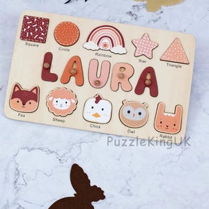 May include: A rectangular wooden name puzzle featuring the name LAURA in large, multi-colored letters with small wooden knobs. Below the name are five animal-shaped puzzle pieces: a fox, sheep, chick, owl, and rabbit, with labels underneath each.