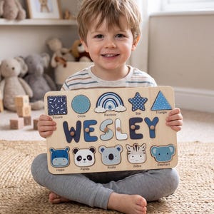 May include: A rectangular wooden puzzle board featuring the name WESLEY in large blue letters, surrounded by cutouts of shapes including a square, circle, rainbow, star, and triangle, plus animal faces labelled hippo, panda, koala, zebra, and elephant.
