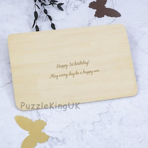 May include: A rectangular light-colored wooden plaque featuring the engraved text Happy 1st birthday! May every day be a happy one. The text PuzzleKingUK is printed in the bottom left corner. Two butterfly cutouts are placed near the plaque.