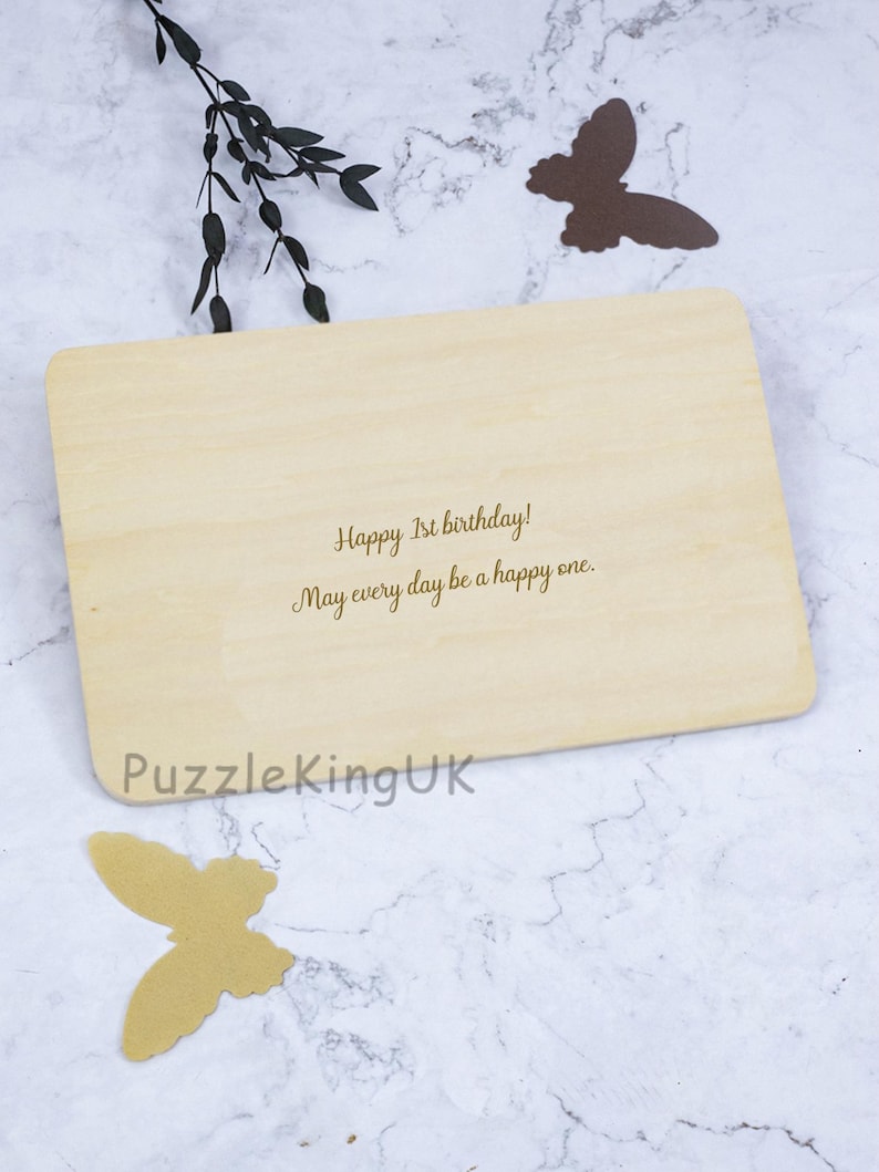May include: A rectangular light-colored wooden plaque featuring the engraved text Happy 1st birthday! May every day be a happy one. The text PuzzleKingUK is printed in the bottom left corner. Two butterfly cutouts are placed near the plaque.