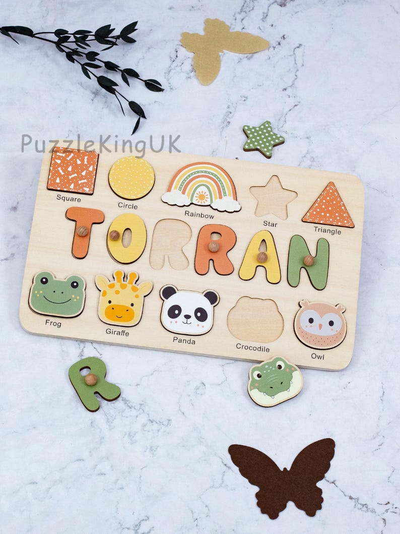 May include: A wooden name puzzle board featuring the name TORRAN in colorful letters with small wooden knobs. The board includes cutouts for shapes like a square, circle, rainbow, star, and triangle, plus animal illustrations of a frog, giraffe, panda, and owl.