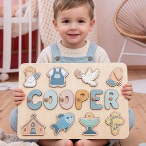 Pode incluir: Um quebra-cabeça retangular de madeira com o nome COOPER em letras grandes de tons pastel com pequenos puxadores de madeira. O tabuleiro inclui recortes com ilustrações de um anjo, uma camisa, uma pomba, uma touca, uma igreja, um peixe, um cálice e uma cruz.