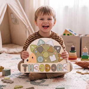 May include: A wooden hedgehog-shaped puzzle board featuring the name THEODORE in large, colorful block letters at the base. The upper section includes five numbered geometric shapes in muted green, brown, and cream tones that fit into the board.