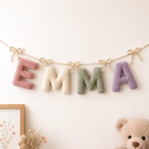 Könnte beinhalten: Eine Häkel-Buchstabengirlande, die den Namen EMMA bildet, mit einzelnen ausgestopften Buchstaben in Altrosa, Creme, Salbeigrün und Lavendel. Jeder Buchstabe hängt an einer naturfarbenen Schnur, die mit kleinen, handgeknüpften Häkelschleifen verziert ist.
