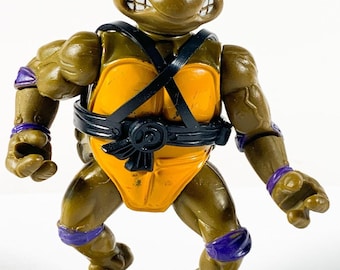 May include: A brown plastic action figure of a turtle character wearing a purple mask, purple wrist and knee bands, and a black harness over an orange chest plate. The figure has a muscular build, a stern facial expression, and clenched hands.