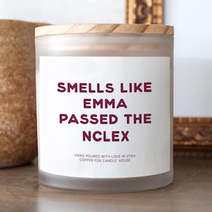May include: A frosted glass candle jar with a light wood lid and a white label featuring the dark red text: SMELLS LIKE EMMA PASSED THE NCLEX, followed by the smaller text: HAND POURED WITH LOVE IN UTAH COPPER FOX CANDLE HOUSE.