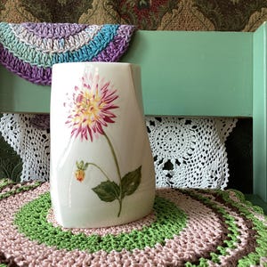 May include: A white ceramic vase with a smooth, slightly asymmetrical cylindrical shape. The front surface features a detailed botanical illustration of a pink and yellow dahlia flower with a green stem, a small bud, and green leaves.