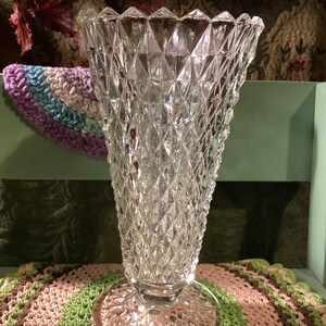 May include: A clear glass vase featuring a prominent diamond-cut pattern across its exterior surface. The vase has a flared, scalloped rim and stands on a circular, textured pedestal base, showcasing a reflective and multifaceted design.