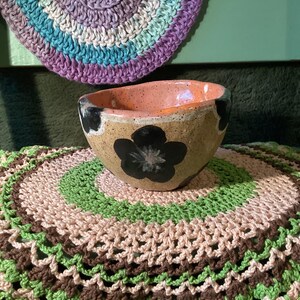 Könnte beinhalten: Eine Keramikschale mit gesprenkelter beiger Außenseite, schwarzen Blumenmotiven und einer glatten, glänzenden pfirsichfarbenen Innenseite. Die Schale hat einen unregelmäßigen, organischen Rand und steht auf einer runden, mehrfarbigen Häkelmatte.