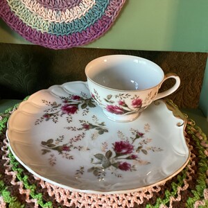 Pode incluir: Uma chávena de chá e um prato a condizer em porcelana branca com um delicado padrão floral de rosas cor-de-rosa e folhas verdes com rebordo dourado. O conjunto é exibido sobre uma superfície de croché texturizada e multicolorida.