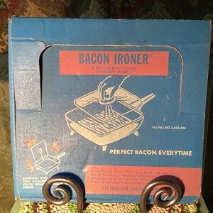 May include: A blue cardboard box for a Bacon Ironer food cooking cover, gourmet model. The front features a graphic of a square press being placed into a skillet, with the text Bacon Ironer, U.S. Pat. No. 3,232,404, and Perfect Bacon Everytime.