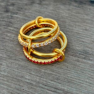 Op de afbeelding: Een stapel van drie gouden ringen. De bovenste ring is een gladde gouden band. De middelste ring is een gouden band bezet met heldere witte stenen. De onderste ring is een gouden band met een rij levendige rode stenen rondom de ring.