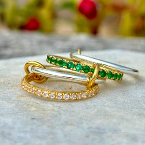 Op de afbeelding: Een verzameling van vier gestapelde ringen, waaronder een gouden band met heldere stenen, een zilveren band, een gouden band met groene stenen en een effen zilveren band. De ringen zijn met elkaar verweven en tonen metalen afwerkingen.