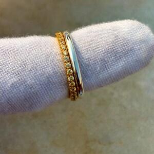 Op de afbeelding: Een tweedelige ringenset bestaande uit een gladde, gepolijste zilverkleurige band en een goudkleurige band met een doorlopende rij kleine, ronde, gele stenen die langs de gehele buitenomtrek van de metalen band zijn gezet.