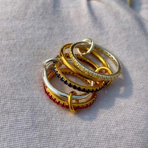 Puede incluir: Un conjunto de cinco anillos metálicos finos entrelazados. El set incluye dos anillos dorados con piedras transparentes, un anillo plateado con piedras azul oscuro, un anillo dorado con piedras azul oscuro y un anillo dorado con piedras rojas.