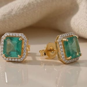 May include: A pair of gold stud earrings featuring a square-cut emerald green centre stone surrounded by a halo of small white pavé crystals. The earrings have a classic post-back closure and a polished gold metal setting that frames the stones.