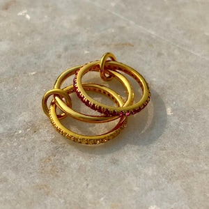 Può includere: Tre anelli dorati intrecciati, ognuno caratterizzato da una fascia sottile. Un anello è incastonato con piccole pietre rosse vivaci, un altro con pietre gialle e il terzo è una fascia dorata lucida e liscia. Gli anelli sono collegati in un gruppo circolare.