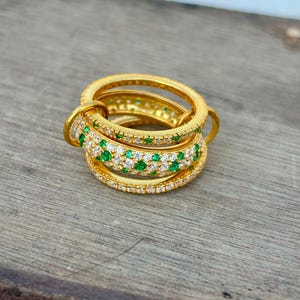 Op de afbeelding: Een goudkleurige ring bestaande uit drie met elkaar verbonden banden. De middelste band is ingelegd met kleine, afwisselend helderwitte en levendig groene steentjes, terwijl de twee buitenste banden van effen goud zijn met een textuur van kraalranden.
