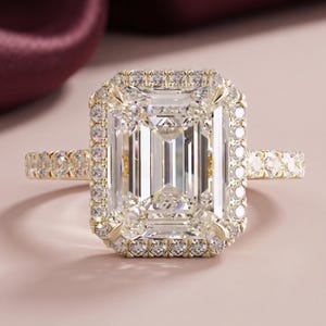 May include: A gold ring featuring a large emerald-cut center stone surrounded by a halo of small round stones. The band is also set with a row of small round stones along the sides, creating a continuous sparkling effect around the finger.