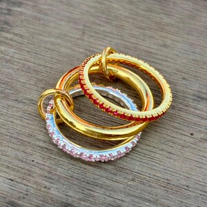 May include: A set of three interlocking rings consisting of one plain gold band, one gold band encrusted with small red stones, and one silver-toned band featuring a row of light pink stones, all linked together to form a single decorative jewellery piece.