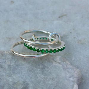 Op de afbeelding: Een set van twee in elkaar grijpende zilveren ringen. De ene ring heeft een gladde, gepolijste band, terwijl de andere ring is bezet met een doorlopende rij kleine, levendige groene steentjes die de hele omtrek van de band sieren.