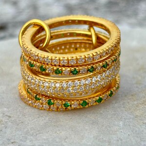 Op de afbeelding: Een stapel van vier goudkleurige ringen verbonden door een kleine gouden lus. De ringen zijn voorzien van afwisselende banden van heldere witte stenen en kleine groene stenen die in het metaal zijn gezet, wat zorgt voor een textuurrijk uiterlijk.