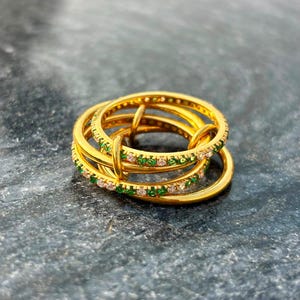 Op de afbeelding: Een goudkleurige ring met een gelaagd ontwerp bestaande uit drie met elkaar verbonden banden. Twee van de banden zijn versierd met een doorlopende rij kleine, afwisselend groene en heldere steentjes, terwijl de derde band van glad metaal is.